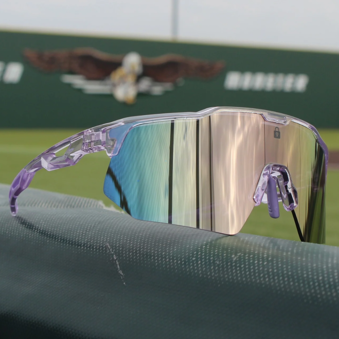 Image of PA10 Polarized Baseball Sunglasses sitting on the dugout featuring a baseball field in the background. With a vibrant reflective mirror lens and pink clear frame the PA10 is ready to hit the field.