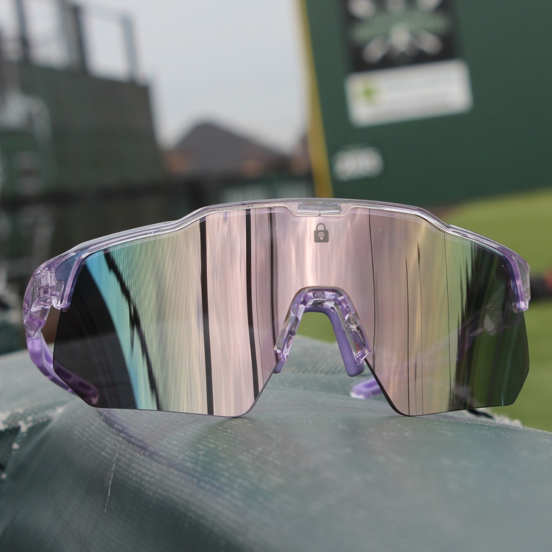 Front view of PA10 Polarized Baseball Sunglasses with a vibrant reflective mirror lens and pink clear frame, resting on the dugout railing with a baseball field in the background, ready for action.