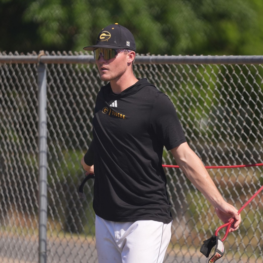 Baseball player stretching with resistance band while wearing sport sunglasses and a team cap during practice.