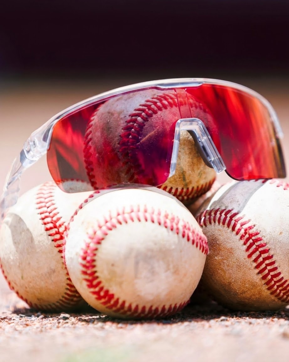 Locked Eyewear Revelation red lens baseball sunglasses in a clear frame resting on game baseballs, showing the non-polarized high-contrast lens built for tracking the ball on sunny fields